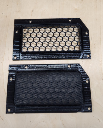 Two rectangular black vent covers on a wooden surface; one with hexagonal grid, one with mesh.